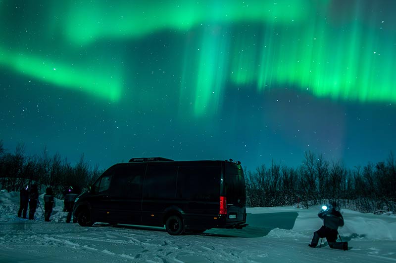 Viajantes observando a aurora boreal ao lado do veículo durante o roteiro pela Noruega